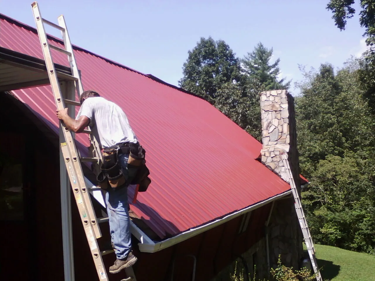 Roofer on a ladder beside a red metal roof and stone chimney — Soffit & Fascia Repair by Roofing Brothers in Eastchester